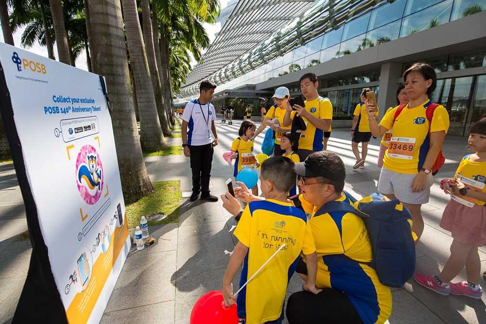 A couple of family and their children gathering around to scan an image of POSB's mascot, Smiley the squirrel, to download the POSB Passion Run for Kids AR Mobile App
