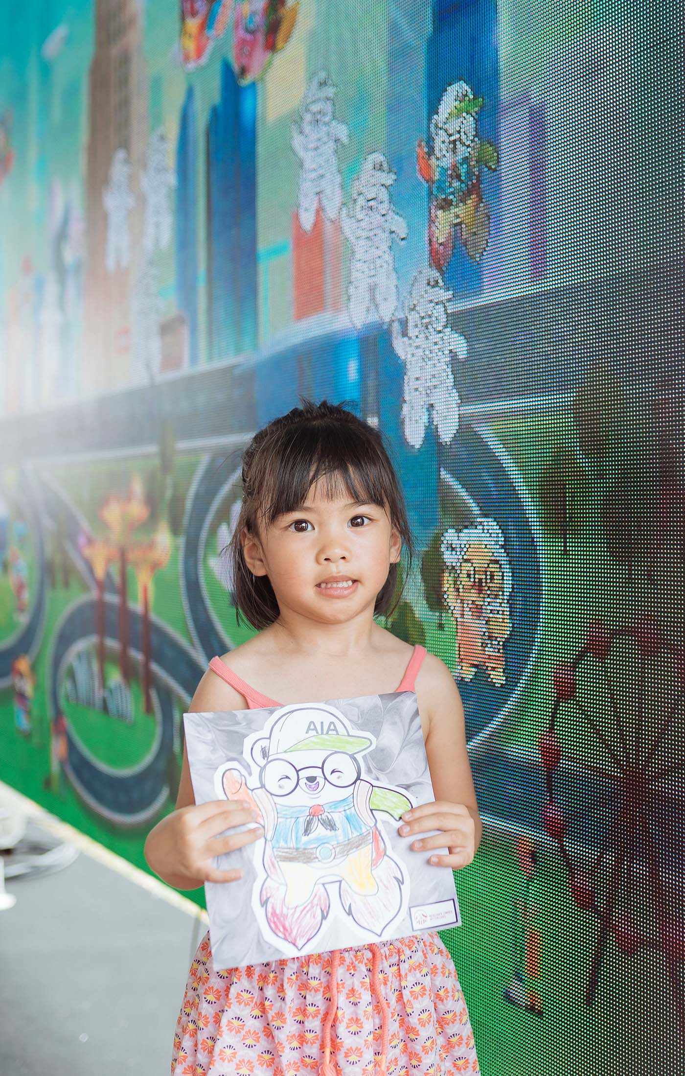 A little girl poses with her art work in front of the AIA Family Fest Coloring Social Wall
